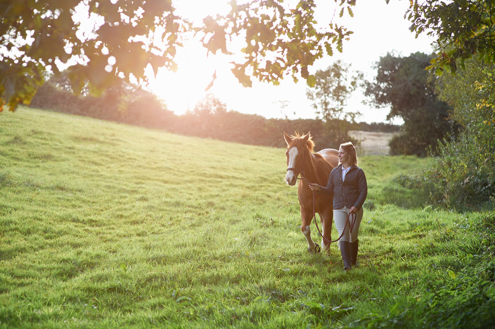 Kvinna promenerar tillsammans med sin bruna häst över en grön äng vid soluppgången